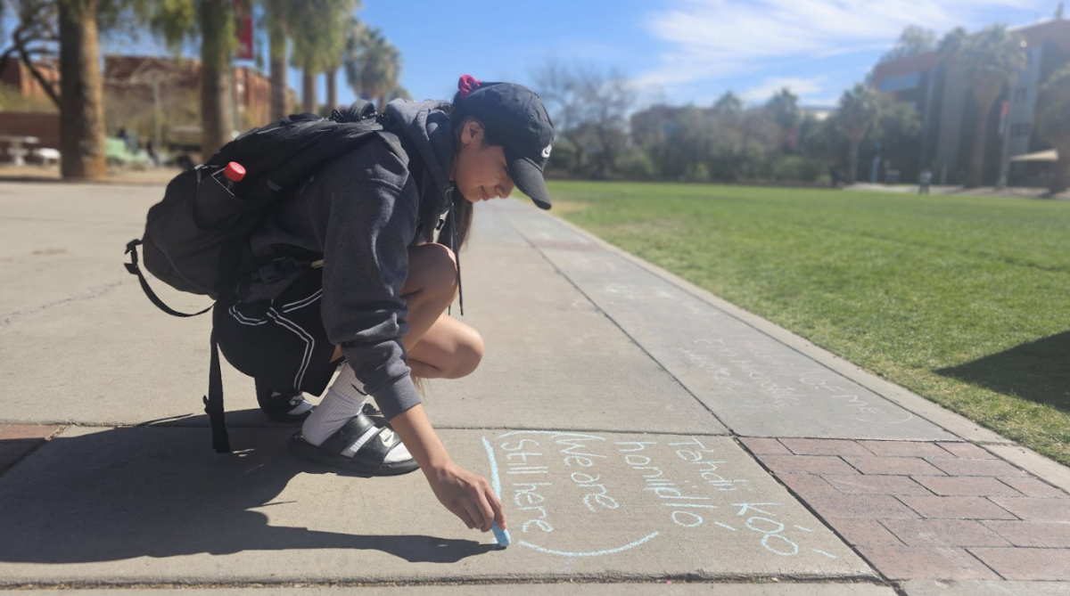 On Mother Language Day, a University of Arizona student makes sure that the world knows that Native American people are still alive. With light blue chalk she writes “we are still here” in Diné and English on the grey asphalt of East University Boulevard. 
